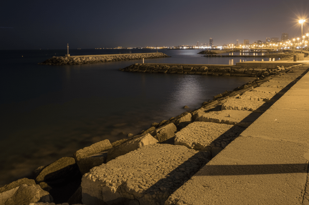 Wide-angle view of illuminated breakwaters and jetties along Barcelona’s Port Olímpic under ambient night lighting, highlighting maritime hazards