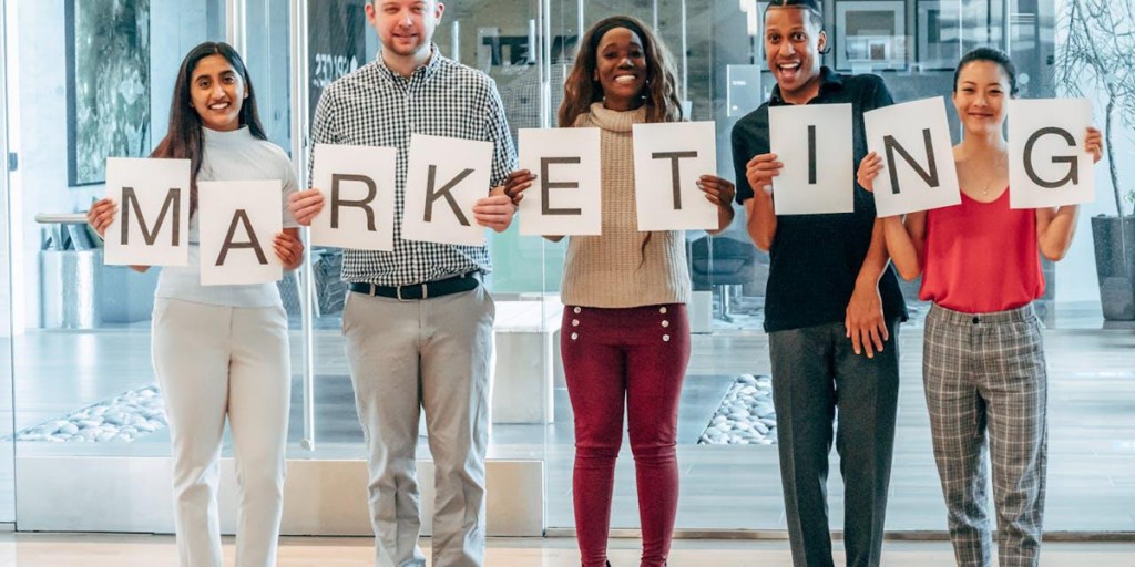 Five people holding up letters printed on paper that spell marketing