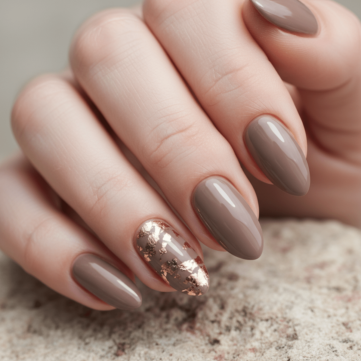 Close-up of mushroom taupe nails with elegant gold leaf accents on a stone background.