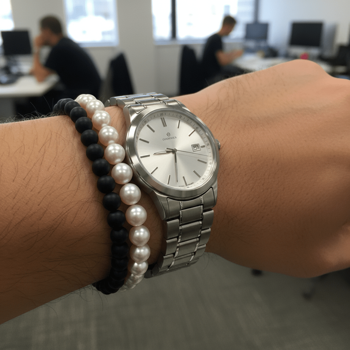 Man's wrist with pearl, steel watch, and matte black beaded bracelets.