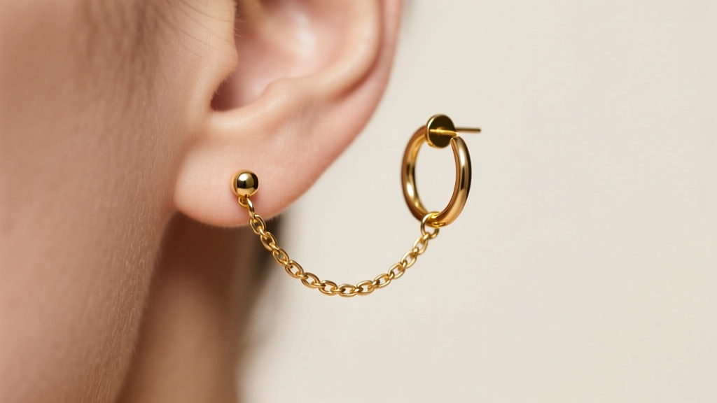 A delicate gold double piercing earrings chain connecting a lower lobe stud to an upper lobe hoop in soft studio light