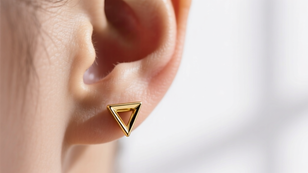 Macro shot of an earlobe wearing a 14k gold equilateral triangle stud pointing down, illuminated by soft, natural window lighting.