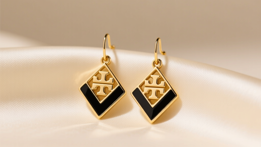 Gold-tone Tori Burch Kira chevron drop earrings with black enamel resting on cream silk fabric under soft, warm studio lighting.