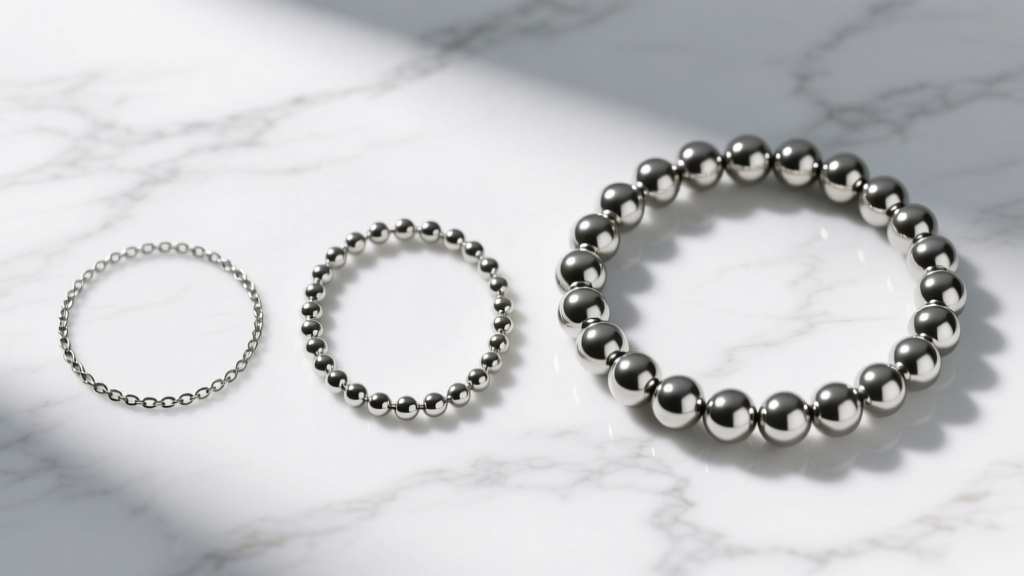 Three steel ball chains on white marble, comparing a thin 2mm spaced chain, a 3mm chain, and a thick 6mm statement bead necklace.