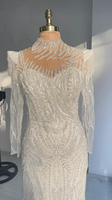 Gaun pengantin putih berbahan renda, model sweetheart neck, lengan panjang, ilusi payet manik-manik, panjang lantai, model mermaid/trumpet, dengan detail rumbai di bahu dan motif bunga.