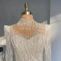 Gaun pengantin putih berbahan renda, model sweetheart neck, lengan panjang, ilusi payet manik-manik, panjang lantai, model mermaid/trumpet, dengan detail rumbai di bahu dan motif bunga.