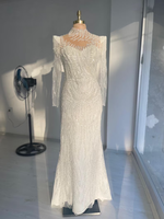 Gaun pengantin putih berbahan renda, model sweetheart neck, lengan panjang, ilusi payet manik-manik, panjang lantai, model mermaid/trumpet, dengan detail rumbai di bahu dan motif bunga.