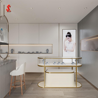 Vitrine d'îlot incurvée ovale pour bijoux, avec garniture en métal doré, dessus en verre, base de rangement laquée crème, unités de comptoir autoportantes