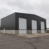 Bâtiment d'atelier préfabriqué en acier gris foncé avec trois portes sectionnelles blanches, base en béton et cour industrielle