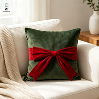 Coussin carré décoratif en velours vert foncé avec nœud rouge, idéal pour les fêtes de Noël et l'ambiance festive à la maison