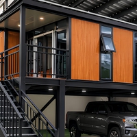 Maison conteneur surélevée à deux étages avec garage, finition bois laminé, structure en acier noir, terrasse vitrée du sol au plafond