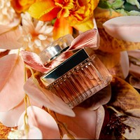 Parfum pour femme en verre côtelé carré avec capuchon argenté, ruban rose, liquide ambré, bouquet doux, pétales romantiques, longue tenue