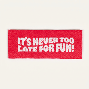 Étiquette de vêtement en polyester tissée et brodée, ronde, rouge, auto-adhésive, avec texte blanc : « Il n'est jamais trop tard pour s'amuser » - Product Image 1