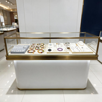 Vitrina de Joyería Rectangular de Vidrio de Lujo con Marco de Metal Dorado, Pedestal Blanco y Luz LED Flotante para Boutique Minorista