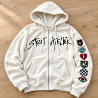 Sweat à capuche oversize crème zippé avec imprimé graphique sur la poitrine, patchs brodés sur les manches, épaules tombantes, style streetwear, personnalisable, vente en gros