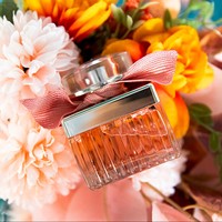 Parfum pour femme en verre côtelé carré avec capuchon argenté, ruban rose, liquide ambré, bouquet doux, pétales romantiques, longue tenue