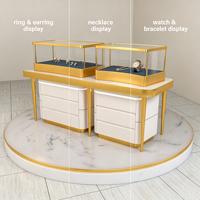 Comptoir de présentation de bijoux compact et combiné, vitrine en verre à cadre fin doré avec base de rangement à tiroirs blancs et plateforme ronde en marbre