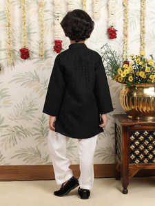 Niño con kurta negra y pijama, en pose de pie lateral, sonriendo, fondo festivo con decoración floral, atuendo tradicional elegante. - Product Image 4