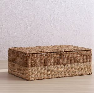 Two-toned <b>rectangular</b> seagrass <b>storage</b> <b>basket</b> with a hinged flat lid, styled on a light wooden floor against a textured wall. - Product Image 1